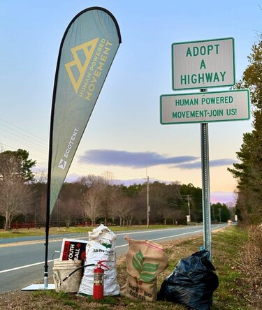 A tear drop flag printed with Ecotent and Human Powered Movement next to an Adopt A Highway sign.